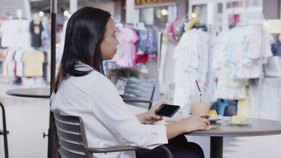 年轻的亚洲女性坐在购物中心的咖啡店前一边吃零食和咖啡一边用智能手机看视频