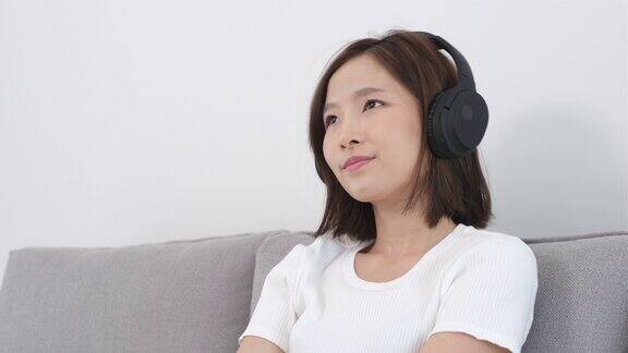 年轻的亚洲女子坐在家里客厅的沙发上戴着耳机听音乐拿着手机