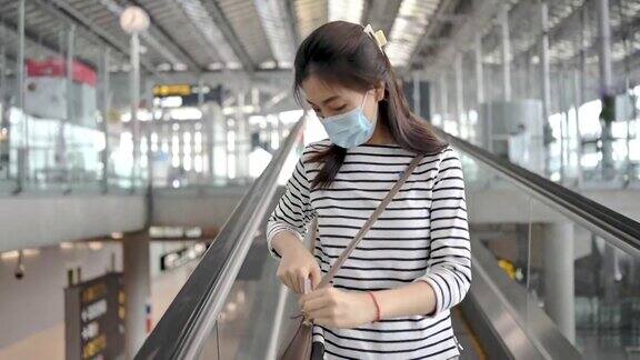 亚洲旅行者女人带着行李戴着口罩在机场候机楼外看站在自动扶梯上去登机口旅行新常态旅游理念