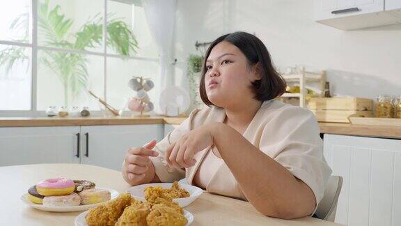 亚洲年轻的胖女人在家厨房里吃不健康的食物迷人的美丽的大码女孩感到饥饿和快乐的咬炸鸡和甜甜甜圈在家里的餐桌上