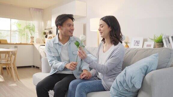亚洲小伙子惊喜地把玫瑰花送给漂亮的女朋友迷人浪漫的新婚夫妇男女在家里的客厅里共度时光在家里庆祝周年纪念
