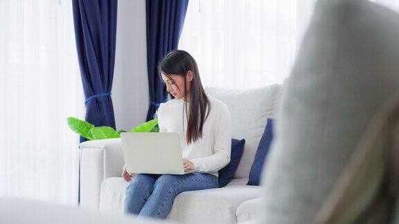 年轻的亚洲女性坐在家里的沙发上使用笔记本电脑