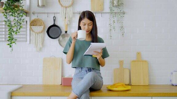 快乐的年轻亚洲女人坐在家里的厨房柜台上女性读书笔记烹饪