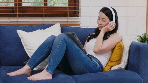 年轻的亚洲女子戴着耳机一边听着数字平板电脑上的音乐一边坐在家里的沙发上生活享受着人们的生活方式