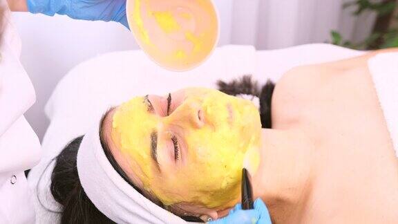 美容师在皮肤护理过程中用刷子覆盖女性面部皮肤的保湿黄色面膜