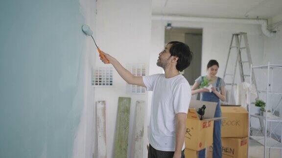 一对年轻的亚洲夫妇在装修他们的新公寓时正在用滚轴用蓝色油漆修补墙壁