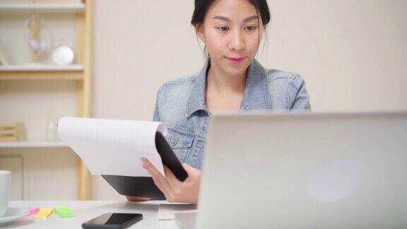 美丽的年轻微笑的亚洲妇女工作笔记本电脑在客厅在家里亚洲商务女性写笔记本文件财务和计算器在家庭办公室享受居家时光
