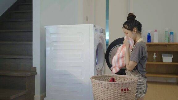 快乐的年轻亚洲妇女打开并从洗衣机取出干净的衣服把衣服放在篮子里疲惫的家庭主妇工作气味