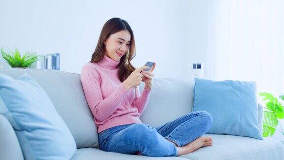 亚洲美女坐在沙发上在家里用手机聊天快乐迷人的年轻女孩在家休闲放松身心享受在客厅用智能手机交流讨论