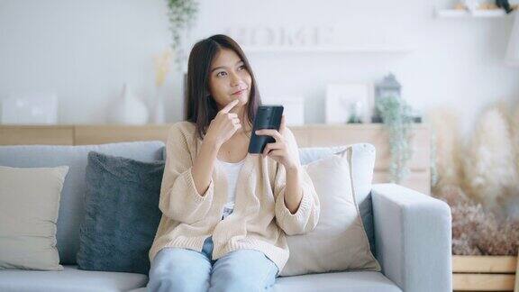 快乐的年轻亚洲女人在家里舒适的沙发上放松在智能手机上输入聊天信息微笑的女孩用手机聊天在浏览器上搜索信息无线互联网网上购物