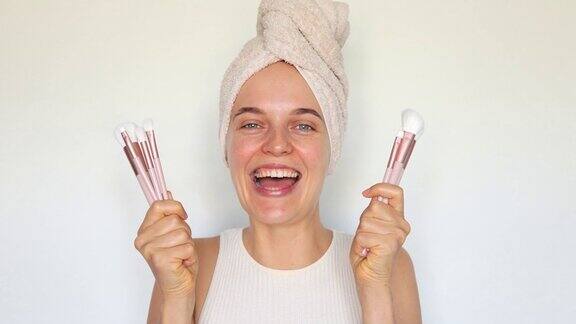 漂亮快乐的女人头上裹着毛巾拿着化妆刷用化妆工具覆盖着脸在白色的背景下心情愉快地做美容手术