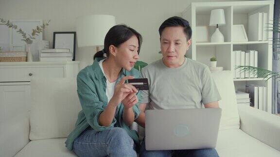 幸福的亚洲夫妇一起坐在家里客厅的沙发上用笔记本电脑购物用信用卡在线支付生活方式花时间在家里