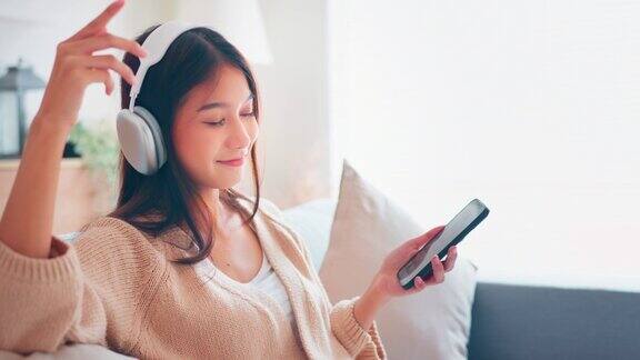 快乐的年轻亚洲女性戴着无线耳机坐在客厅的沙发上用智能手机听音乐享受乐趣女性喜欢在家里听清冷的音乐音频感觉没有压力