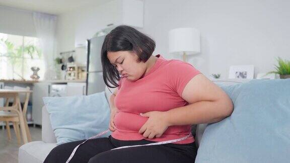 亚洲年轻的胖女人在家里的沙发上用卷尺量腰漂亮的沮丧又不自信的女孩坐在客厅里羞愧地看着肚子想减肥