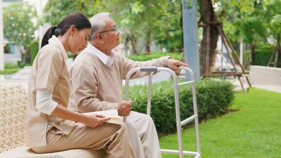 年轻的亚洲女性照顾者照顾者支持老年人老年病人读一本书一个快乐的老人坐在椅子上与步行者在户外公园