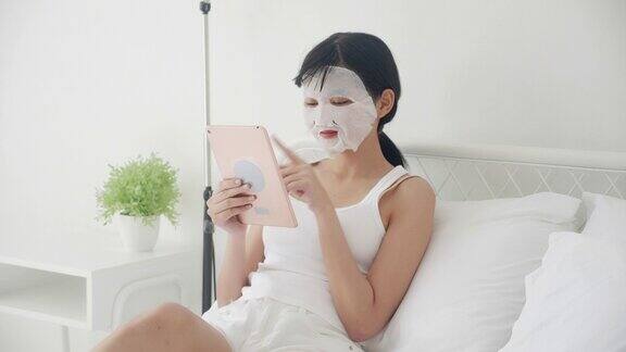 美丽年轻的亚洲妇女与床单面膜坐在床上看平板电脑在卧室美丽的女孩应用面膜的皱纹在家里皮肤护理健康和健康