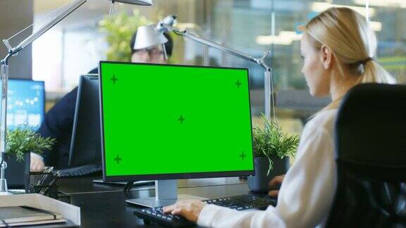 在办公室里女商人在她的办公桌前对着一台模拟的绿色屏幕的个人电脑工作同事走进办公室并在他的办公桌前发生