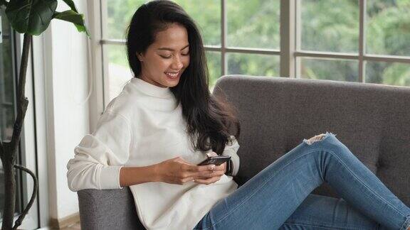 亚洲女人坐在家里的沙发上用手机和朋友聊天社交媒体的生活方式