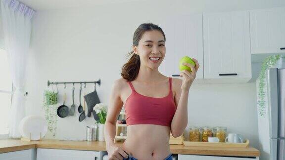 亚洲美女为家里的健康减肥后感到高兴漂亮的女孩手拿青苹果微笑着穿着大牛仔裤在厨房看着镜头健康食品和饮食观念