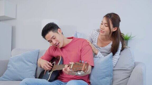 一对亚洲年轻情侣在家里的客厅里一起弹吉他迷人浪漫的新婚男女坐在家里的沙发上玩乐器一起享受周末活动