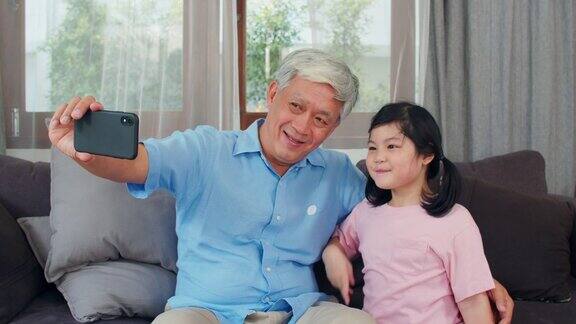 亚洲祖父和孙女在家视频通话中国老爷爷高兴地和年轻的女孩用手机视频通话她的爸爸和妈妈躺在客厅里缓慢的运动