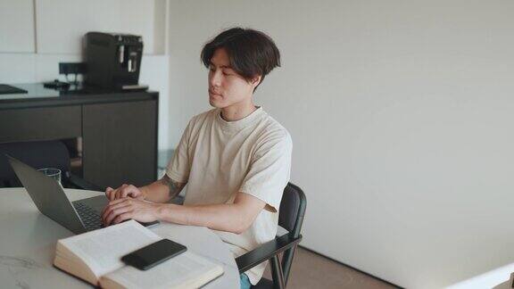沉思的亚洲男子在家里厨房用笔记本电脑工作