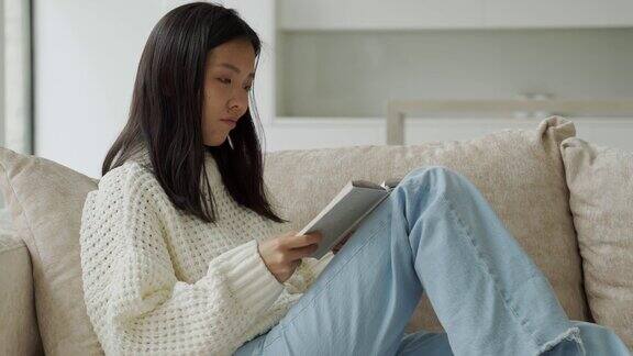 快乐的亚洲女人穿着白色毛衣坐在沙发上看书在家里
