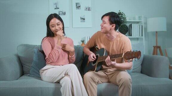 快乐的年轻亚洲夫妇在客厅的沙发上弹吉他唱歌
