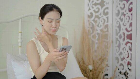 放松的年轻美丽的亚洲女人用面膜坐在床上看智能手机美丽的女孩在家里用面膜护理皱纹皮肤护理健康和保健