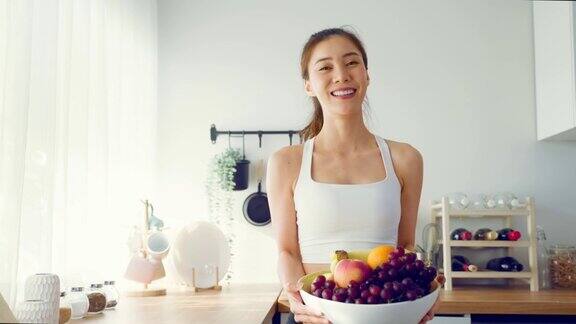 亚洲有吸引力的活跃的女人拿着一碗水果在家里的厨房活跃的运动女孩的肖像在运动服享受吃苹果健康食品为健康锻炼后看在房子里的相机