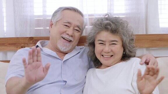 健康的亚洲爷爷坐在客厅的沙发上看着镜头举起双手问候拥抱彼此表达爱意喜气洋洋的老夫妇在屋里微笑