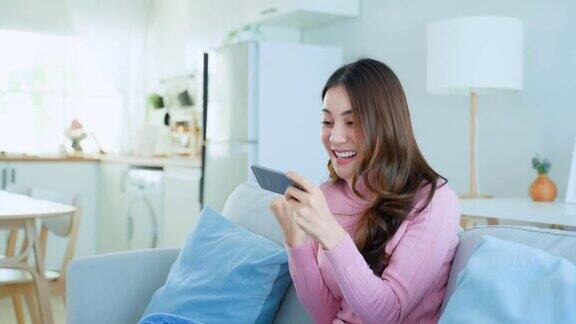 亚洲年轻美女在家里用智能手机玩手机游戏迷人休闲的女孩感到快乐和放松坐在沙发上玩手机触摸屏玩视频在家里享受胜利