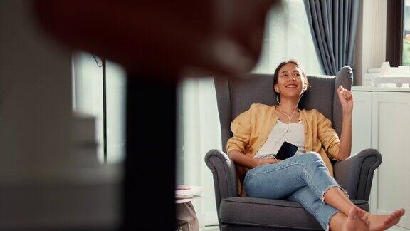 青年亚洲女女孩或大学生戴耳机拿着智能手机坐在沙发椅子上休息chill听音乐唱歌跳舞下班后在家里的客厅