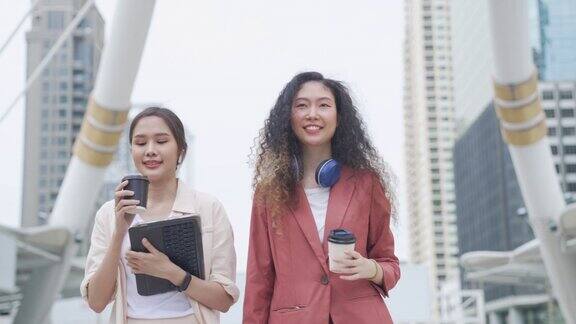 商务往返东南亚女商人拿着咖啡杯和数字平板步行与同事在建筑背景城市工作