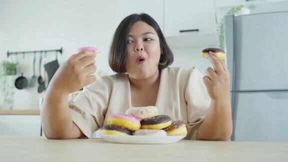亚洲超大身材的女性在家里的厨房里吃不健康的食物年轻漂亮的大码女孩感到饥饿和快乐的咬甜甜甜圈与盘子在餐桌上的家