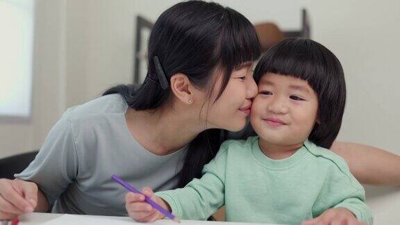 妈妈亲吻儿子教他如何用蜡笔在书上画画或做作业快乐的亚洲男孩与他的母亲在客厅绘画在家里家庭有乐趣