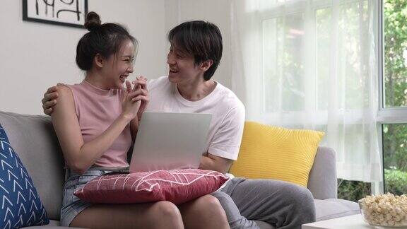 年轻的亚洲夫妇在客厅使用笔记本电脑庆祝他们的成功