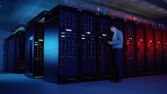 在数据中心:男性IT专家走在一排操作服务器机架上使用笔记本电脑进行维护电信、云计算、人工智能、超级计算机