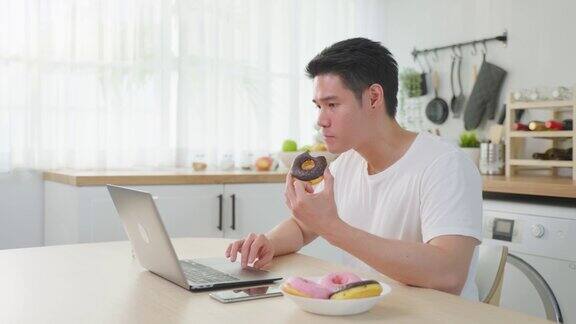 亚洲商人在家工作时吃不健康的垃圾食品有吸引力的英俊年轻男性感到快乐和享受使用笔记本电脑学习网上学习在家里的厨房吃甜甜圈
