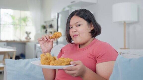 亚洲胖女人的肖像在家里吃不健康的食物漂亮的大码女孩感到饥饿和快乐在客厅的沙发上咬炸鸡微笑看着摄像机