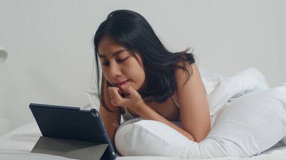 年轻的亚洲女性使用平板电脑检查社交媒体早上在家里醒来后躺在床上感觉快乐的微笑有吸引力的印度女性微笑放松在卧室在家里