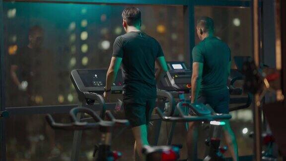 后视图大男子主义的男人力量走跑步机一起在健身房聊天在晚上面对城市的街灯