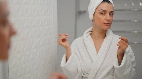 漂亮的年轻多民族女士裹着白色浴巾站在明亮的浴室里用手指按摩脸部快乐的女人对自己的皮肤状况很满意