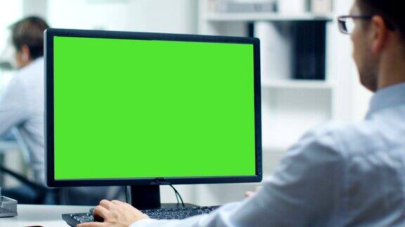 一个商人正在用绿屏的个人电脑工作后台的同事办公室是明亮和现代的