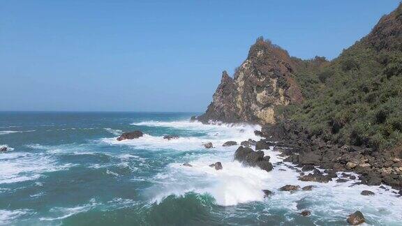 低空飞越岩石海滩海浪拍打岩石溅起的海水无人机飞越岩石海岸线与丘陵景观为背景热带瓦图隆隆海滩印度尼西亚