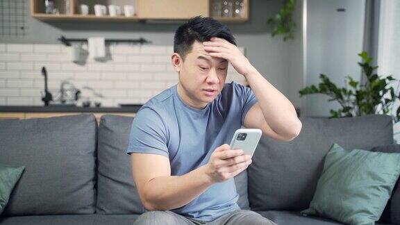 一名亚洲男子坐在家里的沙发上用智能手机看短信室内沮丧地看着智能手机屏幕对收到的信息感到失望