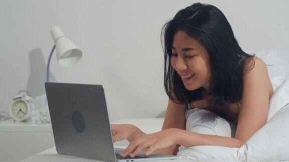年轻的亚洲女人使用笔记本电脑查看社交媒体感到幸福的微笑而躺在床上早上在家里醒来后迷人的泰国女孩微笑放松在卧室在家里