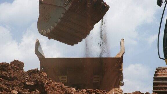 慢动作镜头的一桶挖掘机挖掘捡起泥土并把它倒入一个阳光明媚的建筑工地的自卸卡车的后面
