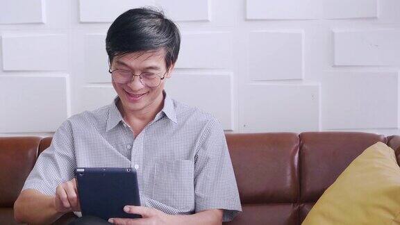 年长的亚洲男子是轻松戴眼镜拿着平板电脑使用互联网检查电子邮件坐在客厅的沙发上愉快地微笑活动生活方式概念家庭科技