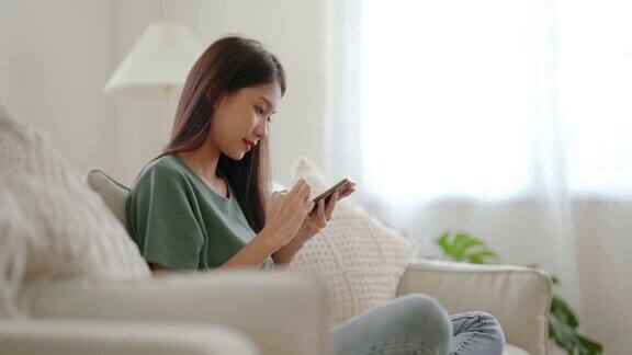快乐的年轻亚洲女性放松在舒适的沙发上在家里用智能手机发短信微笑的女孩使用手机聊天浏览无线互联网上的小工具从家里网上购物